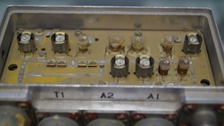 A close-up view of an old electronic circuit board with multiple components including capacitors and resistors. The board is housed in a rectangular metallic casing with labels 'T1', 'A2', and 'A1'. The circuitry appears worn and slightly dusty, suggesting antiquity or frequent usage.