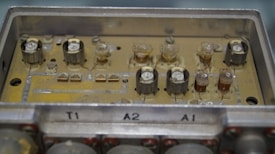 A close-up view of an old electronic circuit board with multiple components including capacitors and resistors. The board is housed in a rectangular metallic casing with labels 'T1', 'A2', and 'A1'. The circuitry appears worn and slightly dusty, suggesting antiquity or frequent usage.