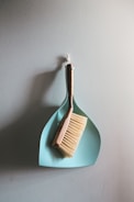 A wooden-handled brush with light bristles is resting on top of a light blue dustpan, both of which are hanging on a textured wall.