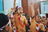 A group of children in school uniforms are animatedly raising their hands, possibly in a classroom setting. They are wearing matching brown uniforms with red and white scarves and headbands.