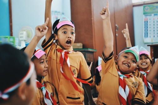 A group of children in school uniforms are animatedly raising their hands, possibly in a classroom setting. They are wearing matching brown uniforms with red and white scarves and headbands.