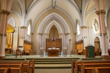 A grand church interior featuring high vaulted ceilings and ornate arches. The altar is centrally located, adorned with intricate woodwork and surrounded by wooden pews. Soft lighting highlights religious symbols, including a cross, and decorative elements like columns and stained-glass windows.