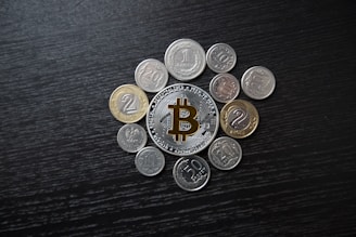A collection of various coins arranged around a central Bitcoin token on a dark wooden surface. The Bitcoin token is prominently featured, with its distinctive logo visible.