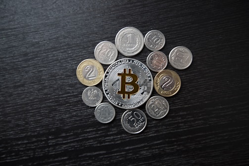 A collection of various coins arranged around a central Bitcoin token on a dark wooden surface. The Bitcoin token is prominently featured, with its distinctive logo visible.