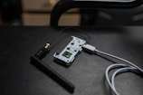 A small electronic circuit board is connected to an antenna and a USB cable. The board features various electronic components and connectors, and it is resting on a dark, textured surface.