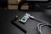 A small electronic circuit board is connected to an antenna and a USB cable. The board features various electronic components and connectors, and it is resting on a dark, textured surface.