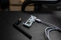 A small electronic circuit board is connected to an antenna and a USB cable. The board features various electronic components and connectors, and it is resting on a dark, textured surface.