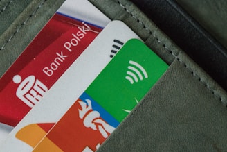 A close-up view of a wallet pocket with several credit cards partially visible. The cards are colorful, featuring logos and symbols including one for contactless payment. The wallet material appears to be leather.