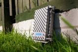 A metallic outdoor electronic enclosure with diagonal ribbing is positioned on a white painted brick surface, surrounded by green grass. A fence in the background casts shadows across the scene, and there is a label with the text 'NEBRA' on the enclosure.