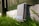 A metallic outdoor electronic enclosure with diagonal ribbing is positioned on a white painted brick surface, surrounded by green grass. A fence in the background casts shadows across the scene, and there is a label with the text 'NEBRA' on the enclosure.