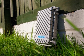 A metallic outdoor electronic enclosure with diagonal ribbing is positioned on a white painted brick surface, surrounded by green grass. A fence in the background casts shadows across the scene, and there is a label with the text 'NEBRA' on the enclosure.