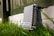 A metallic outdoor electronic enclosure with diagonal ribbing is positioned on a white painted brick surface, surrounded by green grass. A fence in the background casts shadows across the scene, and there is a label with the text 'NEBRA' on the enclosure.