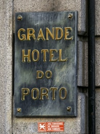 A rectangular metal plaque with raised golden letters displaying the name 'Grande Hotel do Porto' mounted on a stone wall. Below it, a small red sign has text in multiple languages indicating no smoking is allowed.