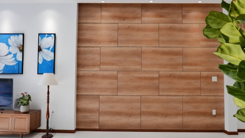 A modern living space featuring a feature wall with wood paneling. To the left, there are two blue and white floral paintings above a wooden cabinet. A tall lamp with a white shade stands next to the cabinet, which has a small plant on top. Part of a large green plant is visible on the right side of the image.