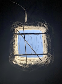 A square window in a dark room with exposed concrete and rebar, allowing sunlight to stream through, illuminating jagged edges. Several wires hang loosely across the opening, with a section of the blue sky visible beyond.