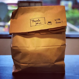 A brown paper bag with a handwritten message that says 'Thank you :) with love!' against an indoor background. The bag is crinkled at the top and sits on a dark surface with a blurred scene visible behind, possibly indicating a window to the outside.