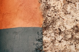 A textured surface showing a contrast between a smooth, painted area on the left and a rough, textured area on the right. The painted section has an orange hue on top and a blue-grey hue beneath, while the textured area appears to be an unpainted, bare section of plaster or concrete with a coarse, uneven finish.
