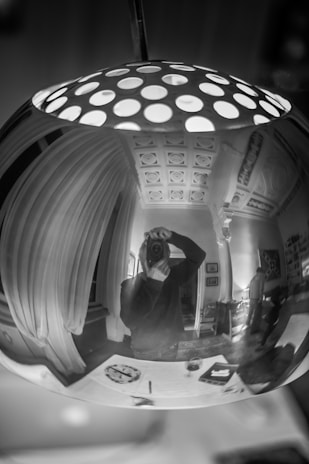 A reflective surface displays a distorted image of a room with ornate ceiling details. A person takes a photograph, capturing themselves in the curved reflection. Various objects, including a book and a glass, rest on a table underneath. The room appears elegantly furnished with curtains and art on the walls.