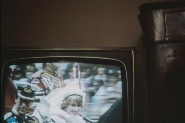 A television screen displaying a ceremonial event with two people in formal attire. The image is slightly blurred and appears to be a broadcast of a parade or public celebration.