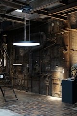An industrial interior with exposed machinery and piping on brick walls. The area is dim with a single glowing light fixture suspended from the ceiling. Metal stairs lead to an upper level and there are various mechanical components and gauges scattered around.