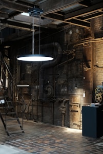 An industrial interior with exposed machinery and piping on brick walls. The area is dim with a single glowing light fixture suspended from the ceiling. Metal stairs lead to an upper level and there are various mechanical components and gauges scattered around.