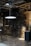 An industrial interior with exposed machinery and piping on brick walls. The area is dim with a single glowing light fixture suspended from the ceiling. Metal stairs lead to an upper level and there are various mechanical components and gauges scattered around.