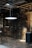 An industrial interior with exposed machinery and piping on brick walls. The area is dim with a single glowing light fixture suspended from the ceiling. Metal stairs lead to an upper level and there are various mechanical components and gauges scattered around.