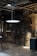 An industrial interior with exposed machinery and piping on brick walls. The area is dim with a single glowing light fixture suspended from the ceiling. Metal stairs lead to an upper level and there are various mechanical components and gauges scattered around.