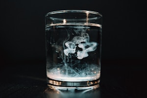 A clear glass filled with water is placed on a dark surface, with a swirl of white smoke or liquid dispersing inside. The background is dark, creating a dramatic contrast with the bright liquid in the glass.