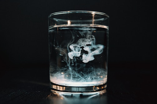 A clear glass filled with water is placed on a dark surface, with a swirl of white smoke or liquid dispersing inside. The background is dark, creating a dramatic contrast with the bright liquid in the glass.