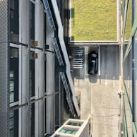 Aerial view of an urban area with a modern building featuring metal and glass construction. The image includes an outdoor staircase along the building's side. Below, a black car is parked in an adjacent driveway, marked with directional arrows. A grassy rectangular section is visible, adding contrast to the industrial setting.