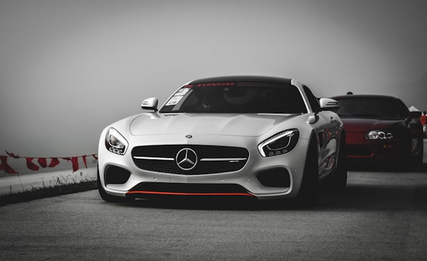 A sleek white sports car prominently displayed on a track, with a large grille and prominent brand emblem. In the background, another dark-colored sports car follows. The overcast sky and blurred background create a dramatic atmosphere.