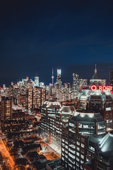 A nighttime cityscape with numerous tall buildings illuminated by artificial lights. In the foreground, a building with a prominent red-lit Rogers sign is visible. The skyline includes a variety of skyscrapers, with the CN Tower distinctly visible among them. The streets below are also lit, indicating a bustling urban environment.