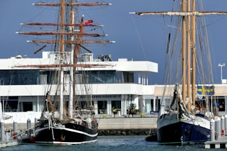 Two tall ships are docked at a marina with a modern white building in the background. The ships have intricate rigging and tall masts, and the water appears calm. The marina is in an urban setting with a mix of nautical and modern elements.