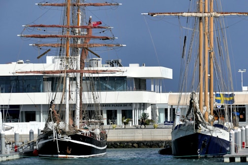 Two tall ships are docked at a marina with a modern white building in the background. The ships have intricate rigging and tall masts, and the water appears calm. The marina is in an urban setting with a mix of nautical and modern elements.
