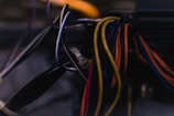 A close-up view of a bundle of various colored electrical wires intertwined, with some in brighter colors like orange, yellow, and blue, set against a dark and indistinct background.