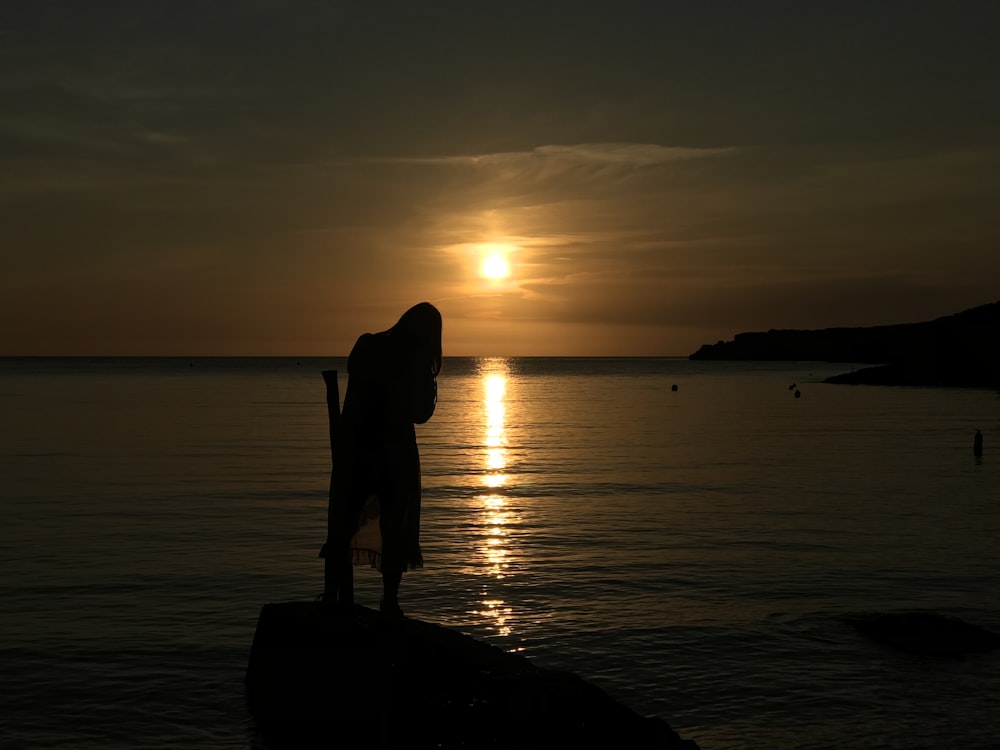 silhouette of woman standing on rock near body of water during sunset