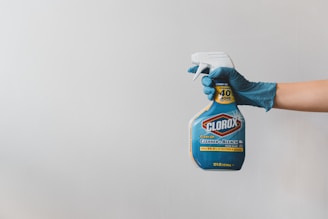 A hand wearing a blue glove holds a spray bottle of Clorox cleaner against a plain white background. The bottle is labeled prominently with blue and yellow colors and features details about its cleaning and disinfecting properties.
