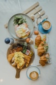 A breakfast or brunch spread on a marble table with two cups of latte art coffee, a plate with an egg and salad, croissants filled with salad and eggs, and a wooden serving board with scrambled eggs, bread, sausages, a hash brown, and some greens.