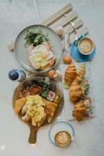 A breakfast or brunch spread on a marble table with two cups of latte art coffee, a plate with an egg and salad, croissants filled with salad and eggs, and a wooden serving board with scrambled eggs, bread, sausages, a hash brown, and some greens.