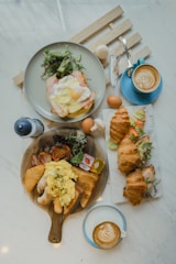 A breakfast or brunch spread on a marble table with two cups of latte art coffee, a plate with an egg and salad, croissants filled with salad and eggs, and a wooden serving board with scrambled eggs, bread, sausages, a hash brown, and some greens.