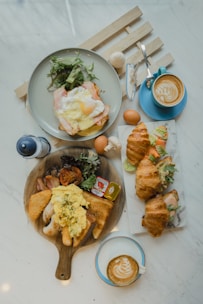 A breakfast or brunch spread on a marble table with two cups of latte art coffee, a plate with an egg and salad, croissants filled with salad and eggs, and a wooden serving board with scrambled eggs, bread, sausages, a hash brown, and some greens.