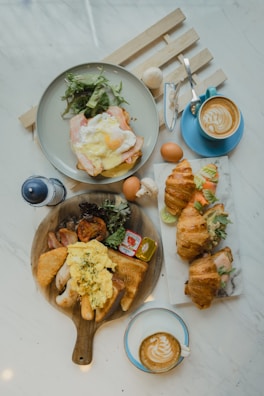 A breakfast or brunch spread on a marble table with two cups of latte art coffee, a plate with an egg and salad, croissants filled with salad and eggs, and a wooden serving board with scrambled eggs, bread, sausages, a hash brown, and some greens.