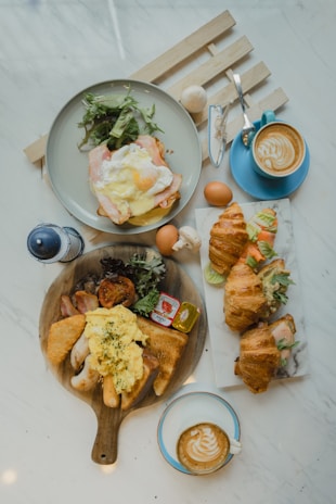 A breakfast or brunch spread on a marble table with two cups of latte art coffee, a plate with an egg and salad, croissants filled with salad and eggs, and a wooden serving board with scrambled eggs, bread, sausages, a hash brown, and some greens.