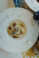 A white plate filled with creamy pasta topped with crispy bacon pieces, garnished with herbs. The plate is set on a marble surface surrounded by scattered uncooked penne pasta and a garlic bulb.