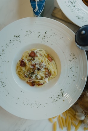 A white plate filled with creamy pasta topped with crispy bacon pieces, garnished with herbs. The plate is set on a marble surface surrounded by scattered uncooked penne pasta and a garlic bulb.