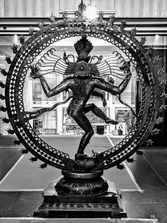 A detailed and intricate bronze statue of the Hindu deity Shiva depicted in the Nataraja pose, surrounded by a circular ring of flames. The statue stands on a pedestal, with one leg raised and the other resting on a small figure, symbolizing the triumph over ignorance. The sculpture is set in an indoor or exhibition space, with a backdrop of architectural elements and other artworks.