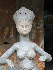 A clay sculpture of a woman with a serene expression and intricate facial features, set against a textured, earthy background. The sculpture's body and face are finely detailed, with a crown-like adornment on the head.