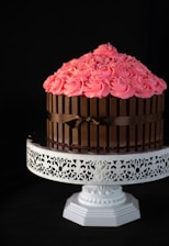 A beautifully decorated cake is placed on an ornate white cake stand. The cake features bright pink frosting swirls adorned with colorful sprinkles on top. Kit Kat chocolate bars surround the sides of the cake, secured with a brown ribbon.