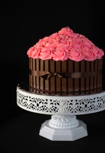 A beautifully decorated cake is placed on an ornate white cake stand. The cake features bright pink frosting swirls adorned with colorful sprinkles on top. Kit Kat chocolate bars surround the sides of the cake, secured with a brown ribbon.